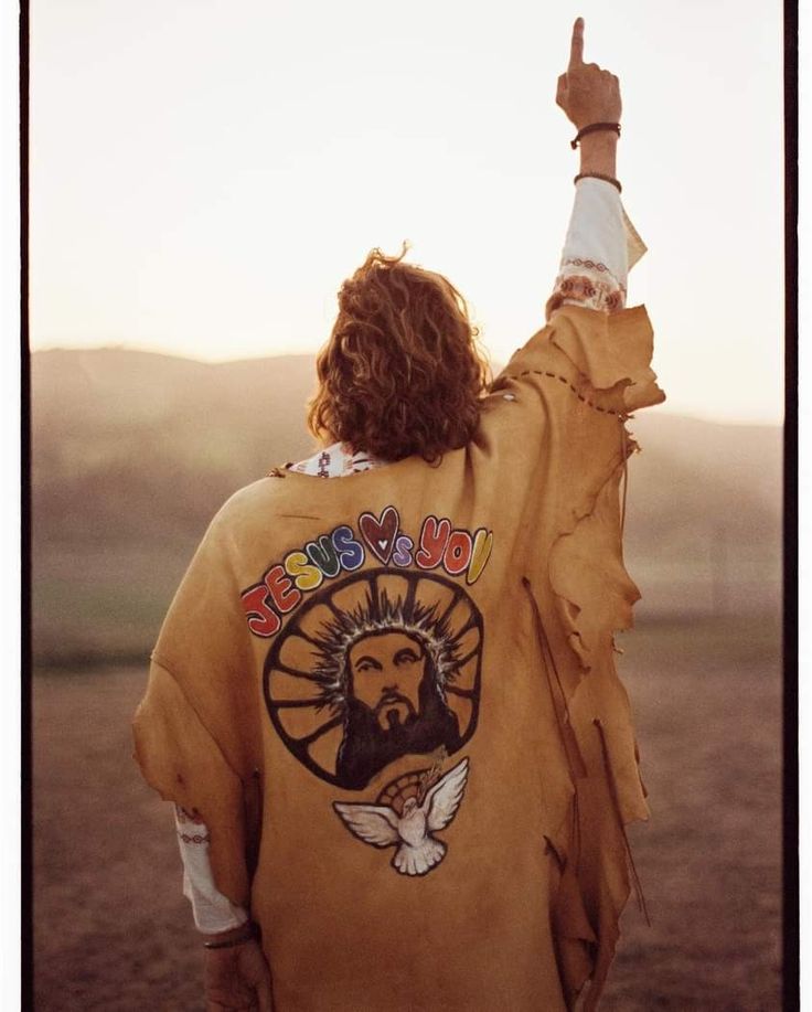 a man with long hair wearing a brown jacket and pointing to something in the distance