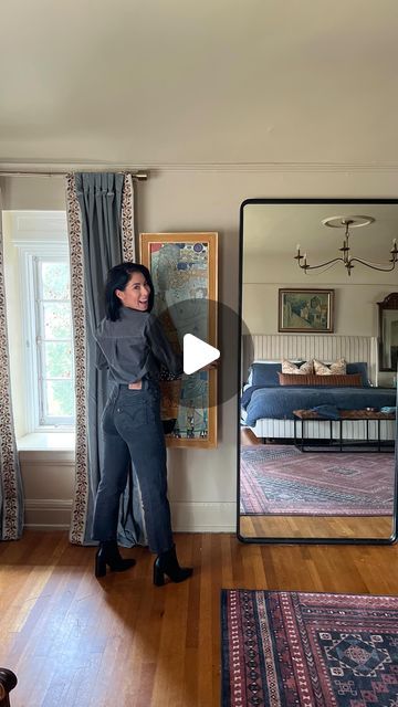 a woman standing in front of a large mirror next to a window with curtains on it