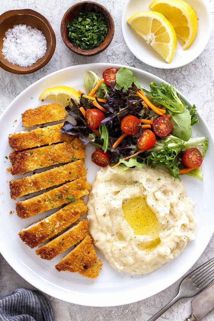 a white plate topped with meat, mashed potatoes and salad next to lemon wedges