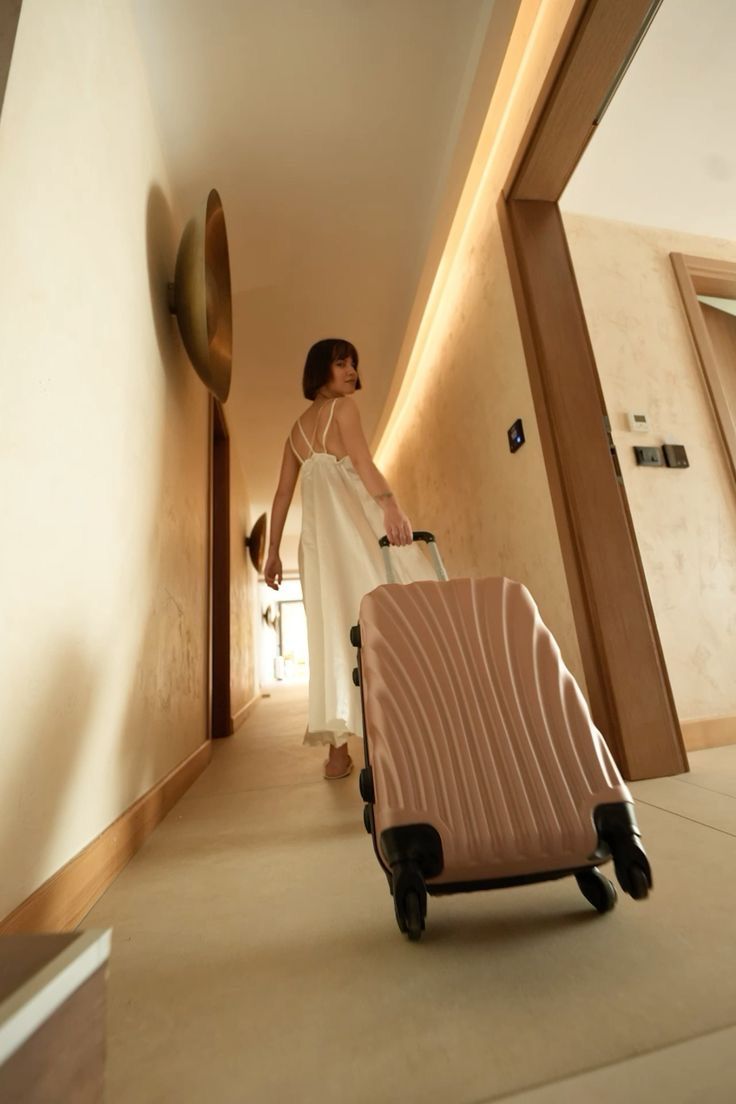 a woman in a white dress pulling a pink suitcase down a hallway with a clock on the wall