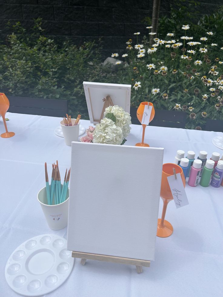 an easel is set up on a table with flowers and paintbrushes in it