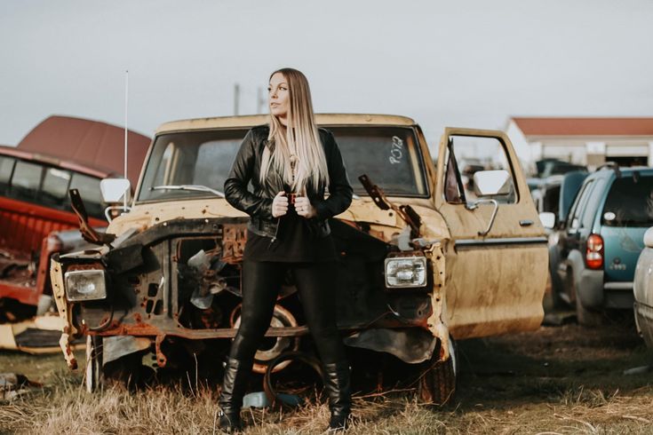 a woman standing next to an old truck in a field with other cars behind her