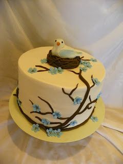 a white cake with blue flowers and a bird sitting on the tree branch in front of it