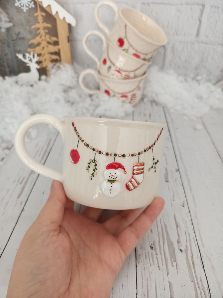 a hand holding a white coffee cup with christmas decorations on it and two mugs in the background