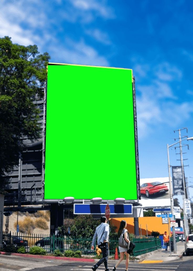 two people walking across the street in front of a large green screen on a building