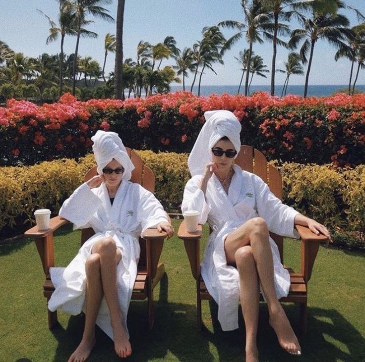 two women in white robes sitting on lawn chairs with palm trees and flowers behind them