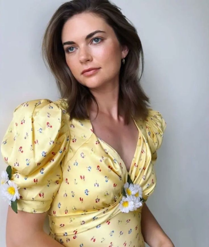 a woman in a yellow dress with flowers on her collared neck and shoulders is posing for the camera