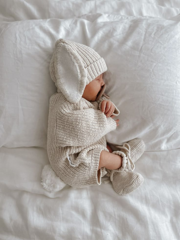 a baby is sleeping on a bed with white sheets and pillows, wearing a knitted outfit