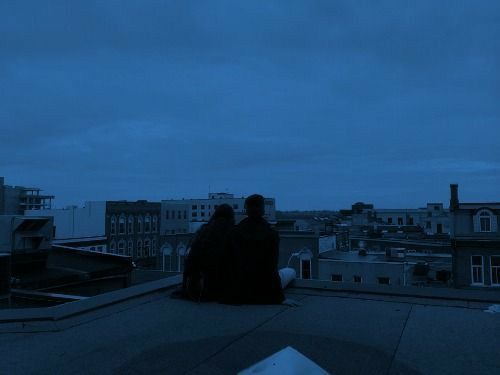 two people sitting on top of a roof looking at the night sky with buildings in the background