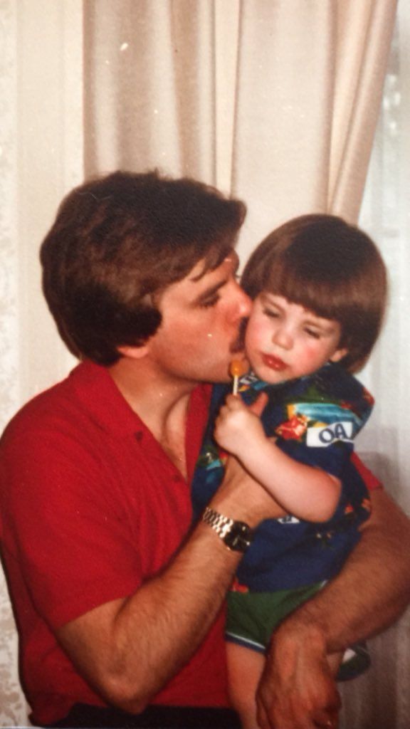 a man holding a little boy in his arms and kissing him on the cheek while sitting next to a window