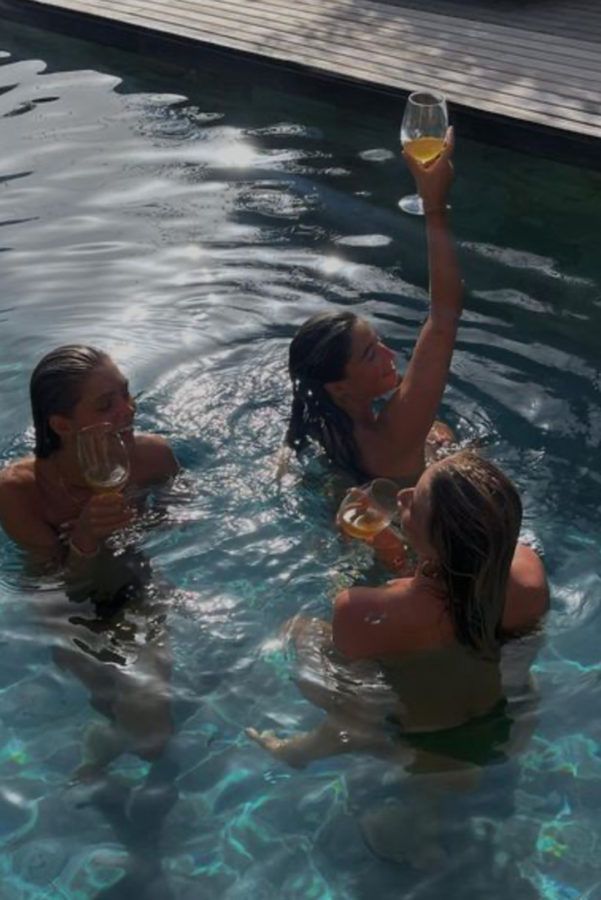 three women in a pool drinking wine and toasting with their hands up to the sky