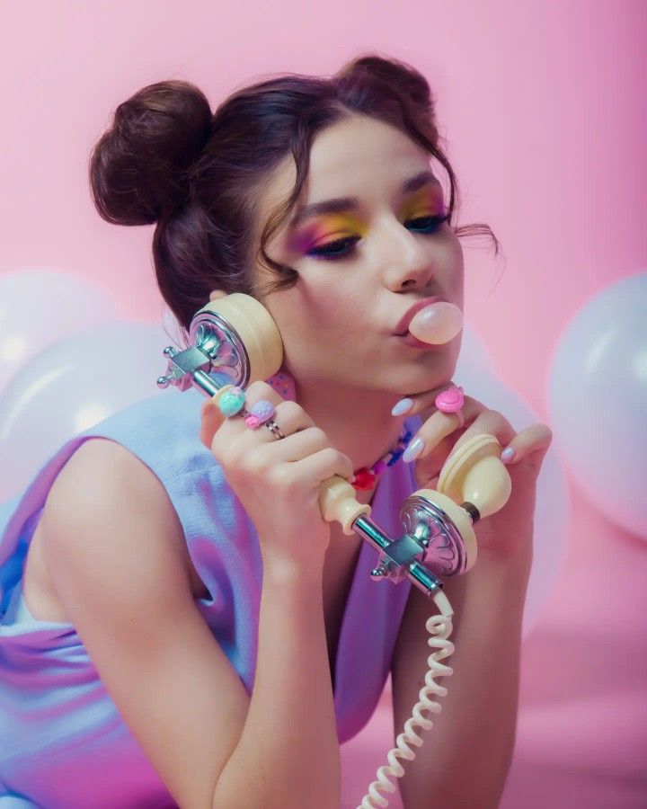 a woman holding a phone up to her face while wearing pink and blue makeup, with balloons in the background