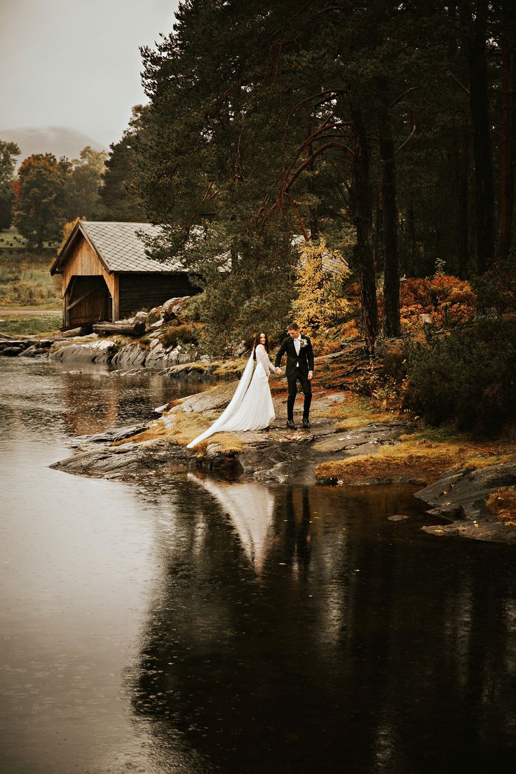 a bride and groom standing on the shore of a lake in front of a cabin