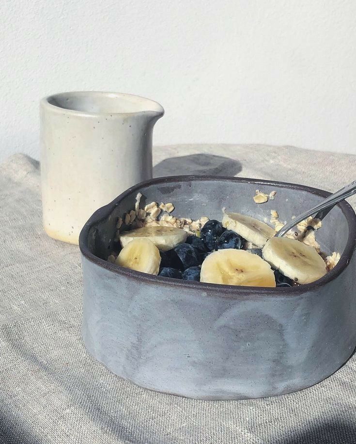 a bowl of cereal with bananas and blueberries in it next to a mug on a table