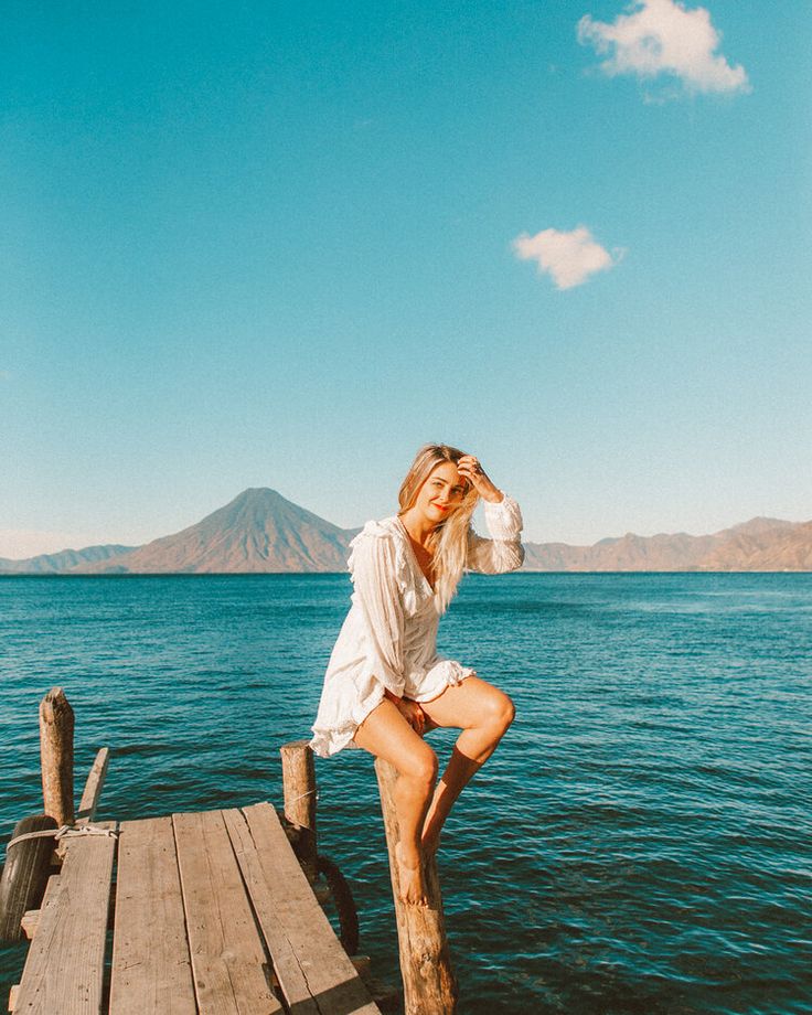 a woman sitting on top of a wooden dock next to the ocean with mountains in the background