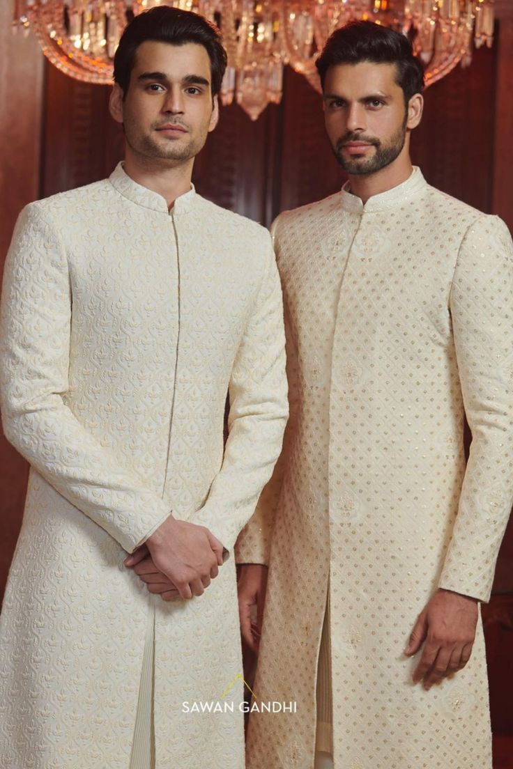 two men standing next to each other wearing white outfits and tuxedos with chandeliers in the background