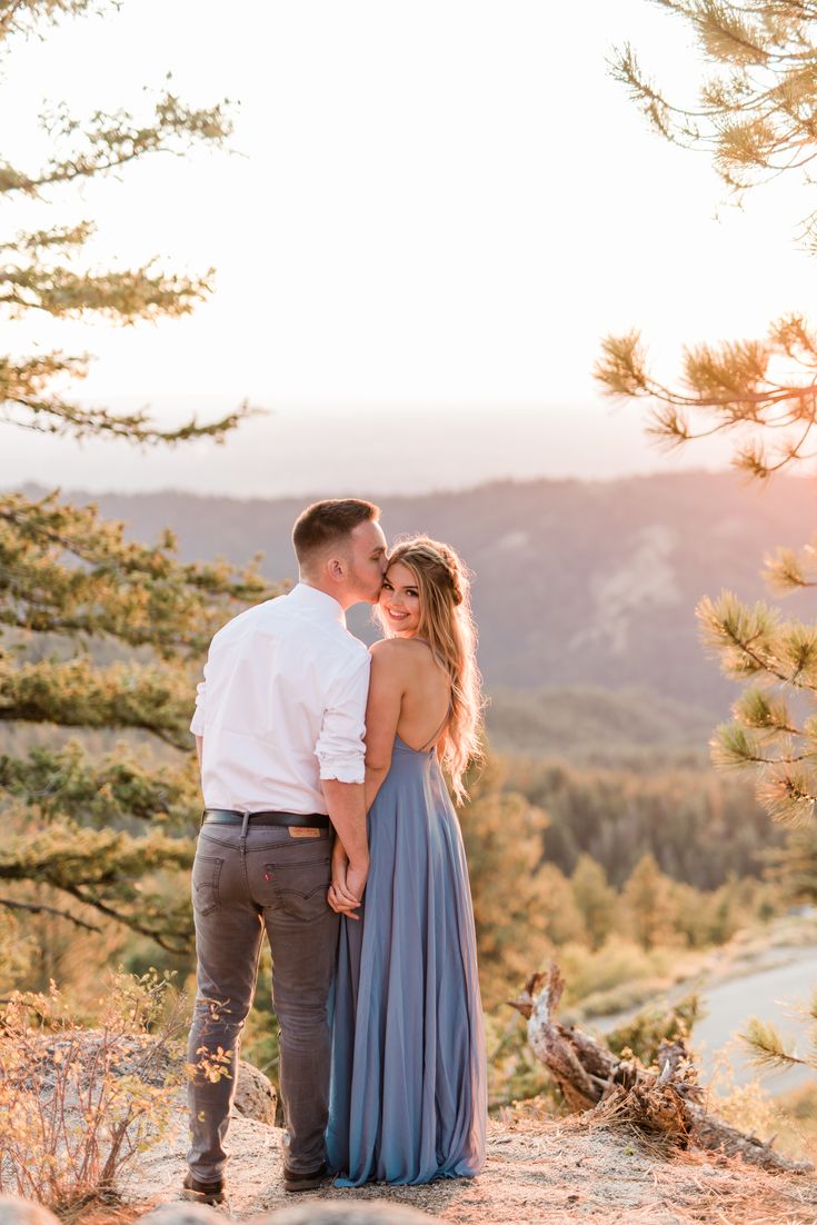 an engaged couple standing on top of a mountain at sunset with the sun setting behind them