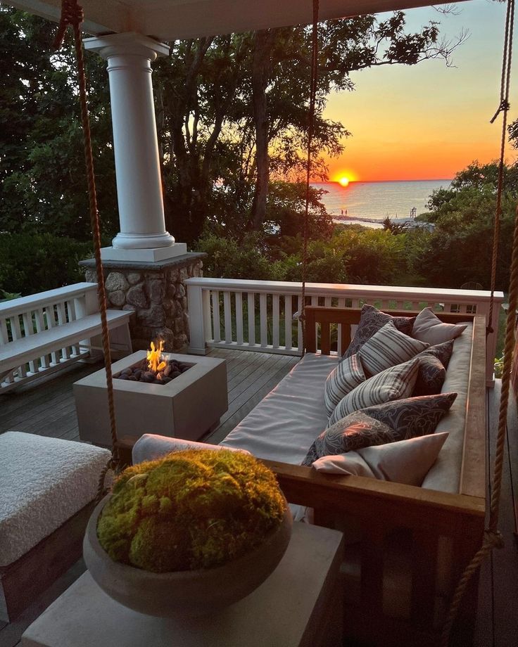 a porch swing with pillows on it and a fire pit in the background at sunset