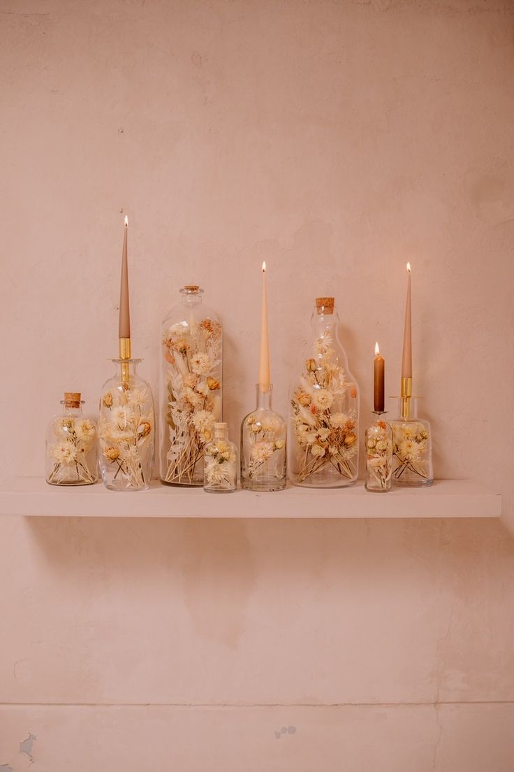 candles and flowers in glass bottles on a shelf next to other vases filled with dried flowers