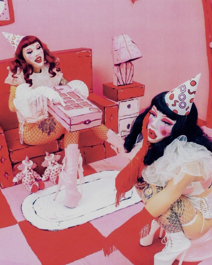 two women dressed up in costumes and hats sitting on a bed with pink walls behind them