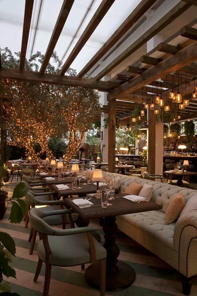 an outdoor restaurant with tables and couches, lights hanging from the ceiling and potted trees in the background