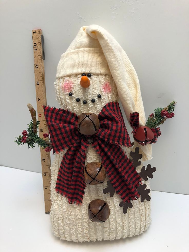 a knitted snowman holding a ruler and wearing a red plaid hat with bells