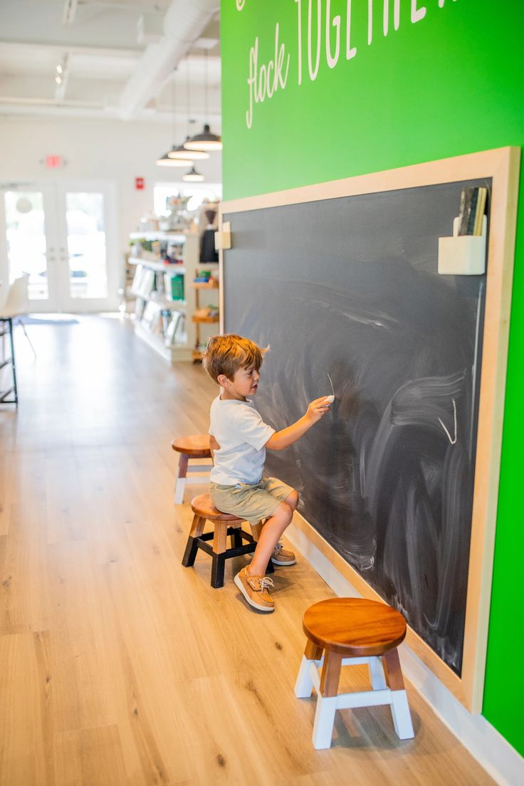 a young boy writing on a blackboard in the middle of a room with wooden stools