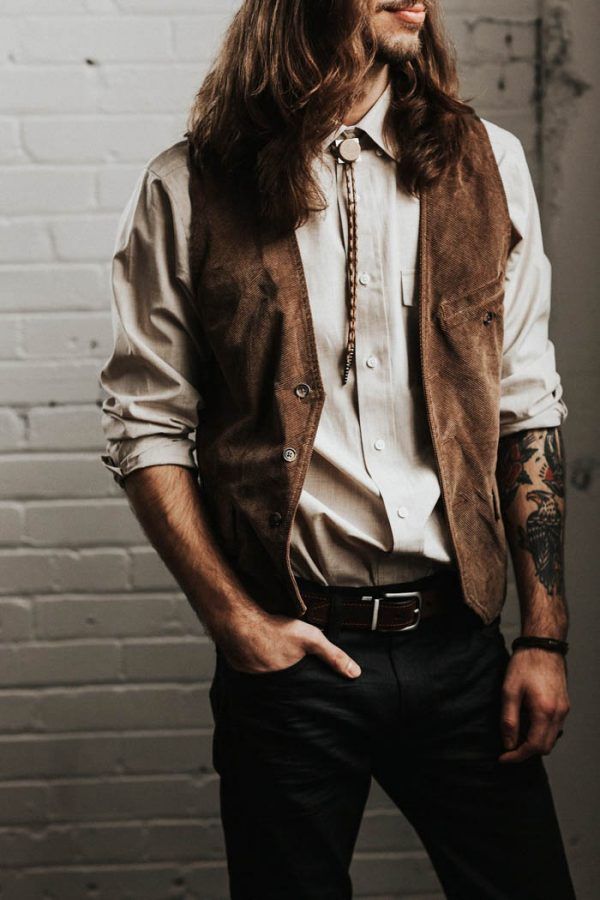 a man with long hair wearing a brown vest and white shirt is standing in front of a brick wall