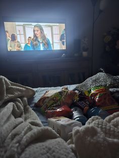 a person laying in bed watching tv with food on the bed next to them and an open bag of chips