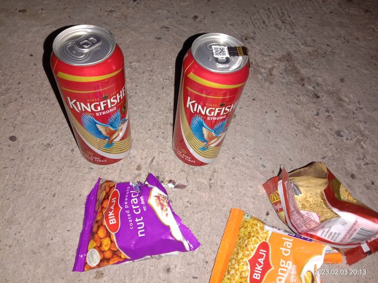 two cans of kingfisher are sitting on the ground next to chips and candy