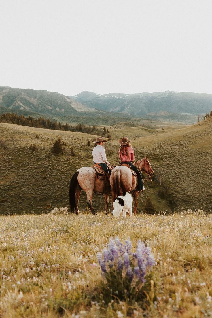 two people riding horses with a dog in the grass and mountains in the back ground