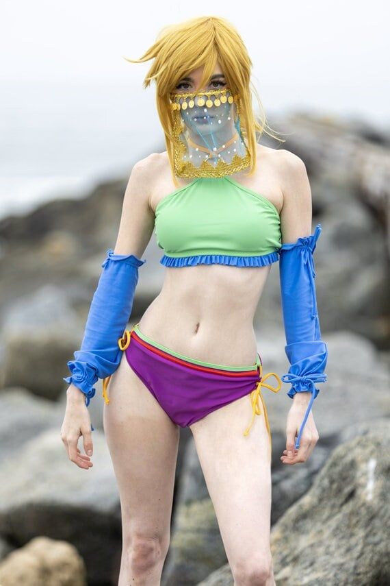 Vasaaq! This bikini swimsuit was inspired by Gerudo Link from Breath of the Wild. Sneak into the desert town by looking the part: the swim wear halter top is lime green with blue ruffles and a gold ring, and ties at the neck with an adjustable back. Cups are sewn into the top. The purple bottom has red and green detail, and ties at the sides with adjustable gold drawstrings. The blue sleeves add extra desert flair. Made from comfortable nylon spandex and ready for use as swimwear. Machine washable in cold water. Look adorable in the desert heat.     Three piece swimwear: top, bottom, sleeves.     Top has cups sewn in.     Adjustable back.     Bottoms have adjustable sides.     Sleeves have elastic ties.     Made from nylon spandex.     Use for swimming and water activities (pool, beach, et Totk Cosplay, Botw Gerudo, Gerudo Link, Desert Town, Swimsuit Cosplay, Purple Bottom, Green Swimwear, Green With Blue, Blue Mask