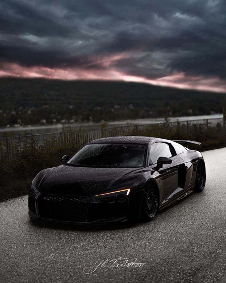 a black sports car driving down a road under a dark sky with clouds in the background