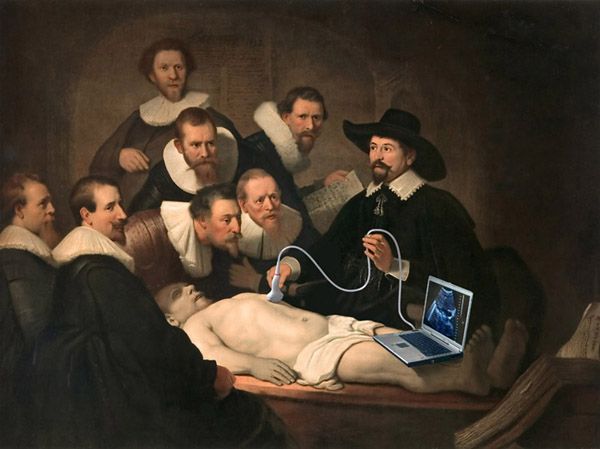 a group of men standing around a man laying on top of a bed with a laptop