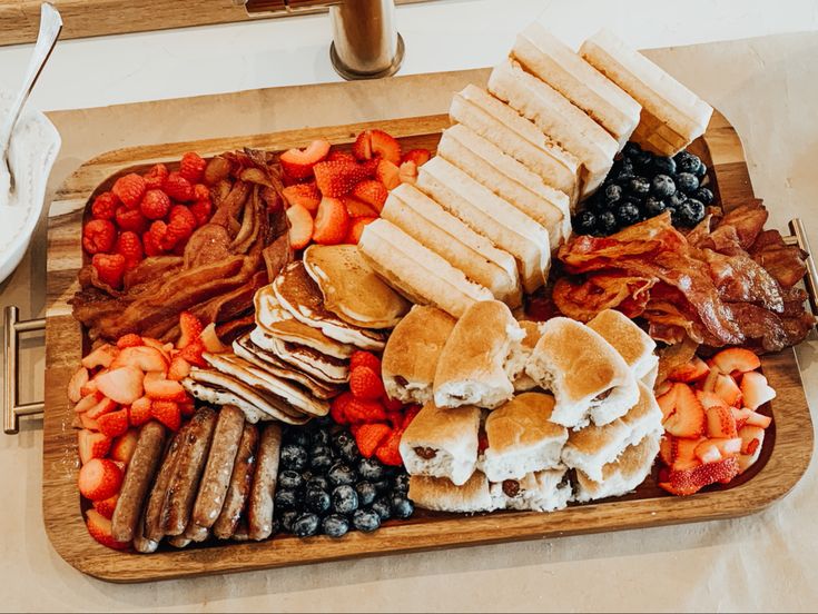 a wooden platter filled with pancakes, bacon, blueberries, strawberries and other breakfast foods