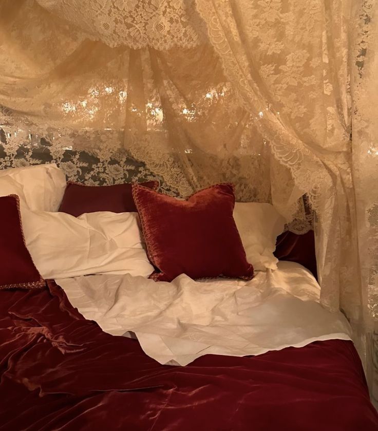 an unmade bed with red pillows and white sheets in a room that has lace curtains on the ceiling