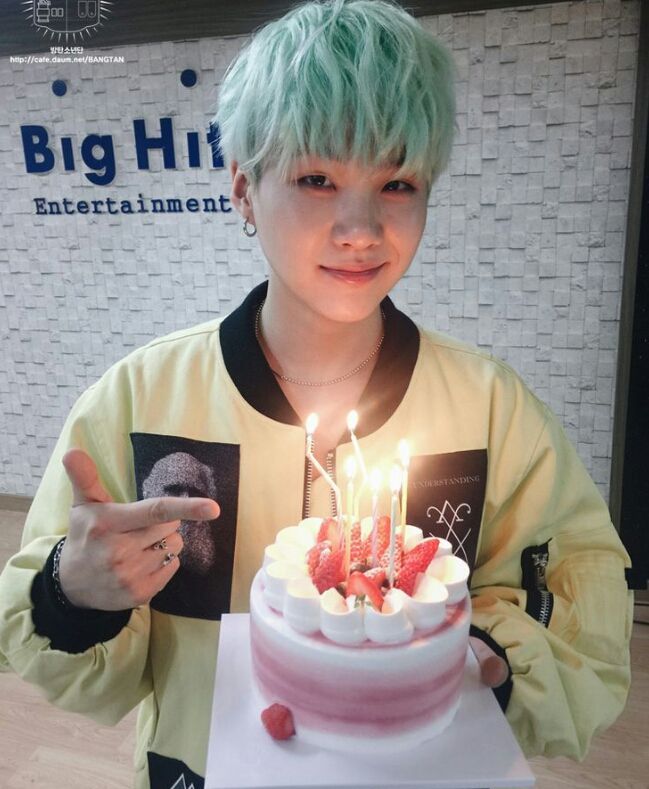 a boy with green hair is holding a birthday cake and pointing at the candles on it