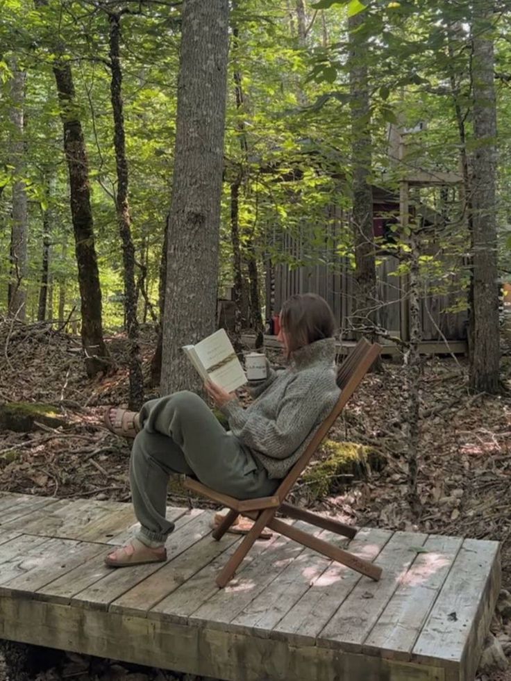 a woman sitting in a chair on top of a wooden platform reading a book outside