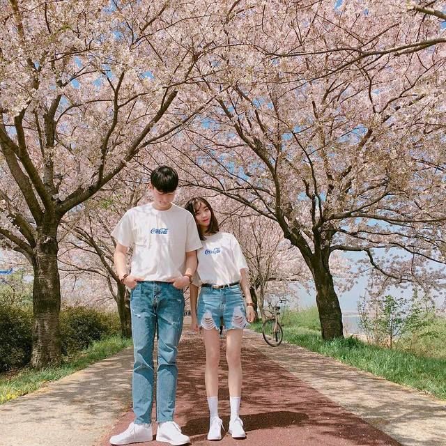 a man and woman walking down a dirt road next to trees with blossoming flowers