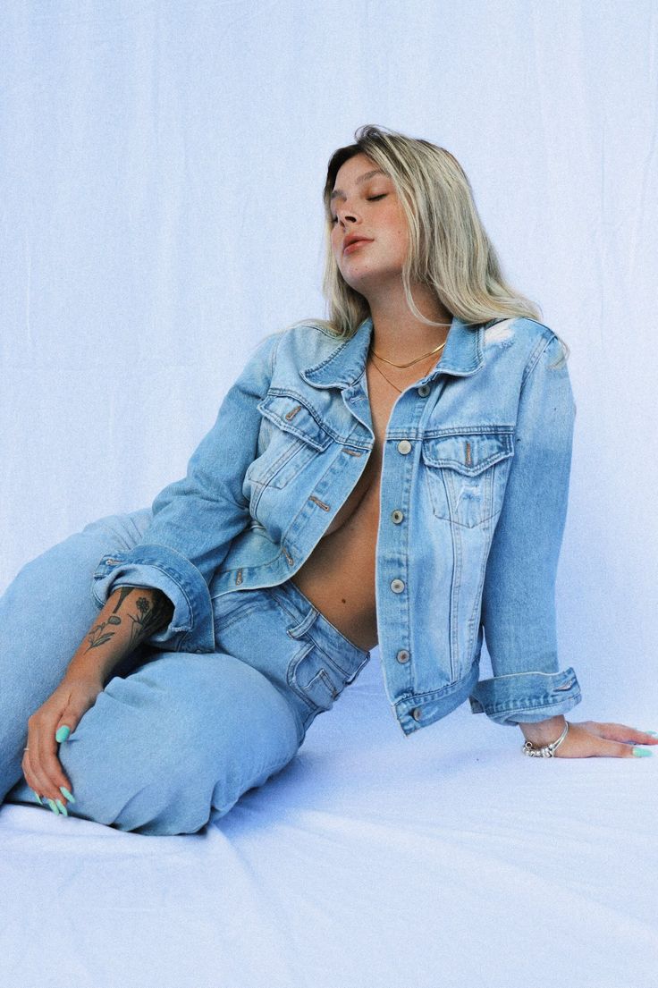 a woman sitting on the ground wearing a jean jacket and blue jeans with her hands in her pockets