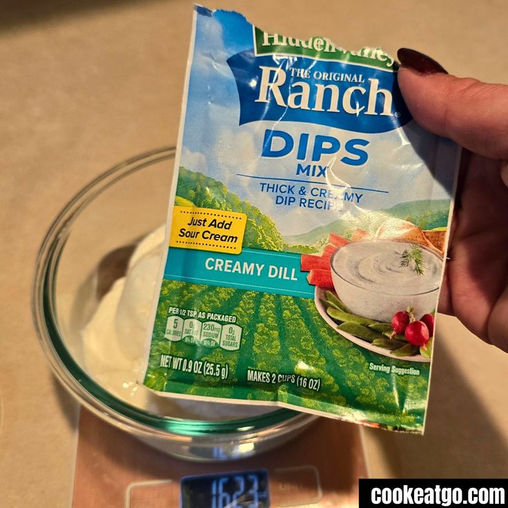 a person holding up a packet of ranch dips in front of a glass bowl