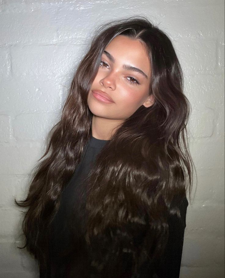 a woman with long brown hair standing in front of a white wall and looking at the camera