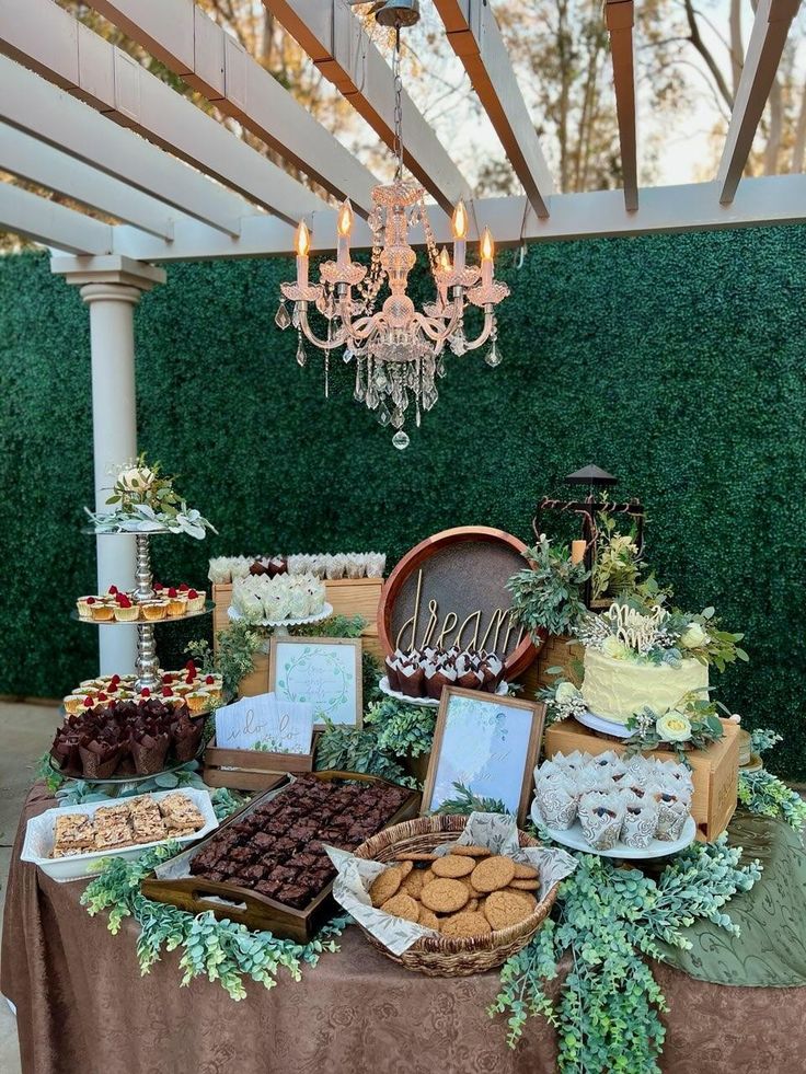 a table topped with lots of desserts under a chandelier covered in greenery