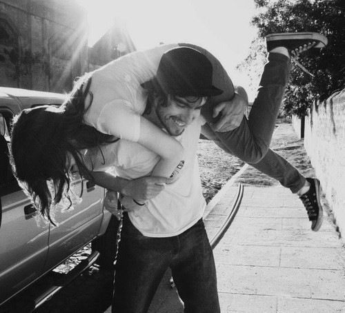 a man carrying a woman on his back while standing next to a parked car in front of a building