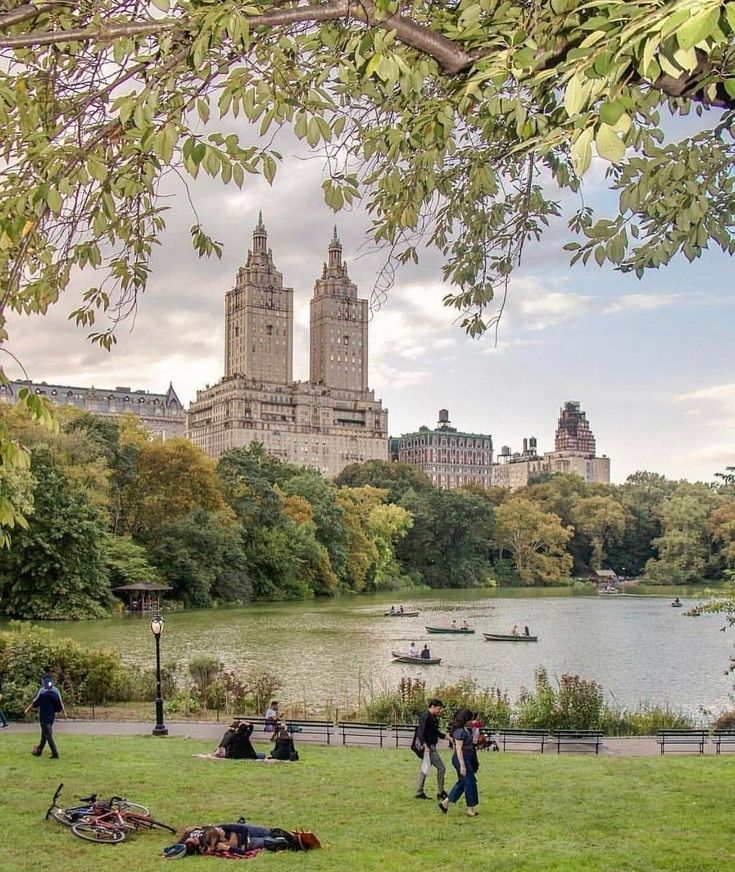 people are walking around in the park by the water and buildings on the other side