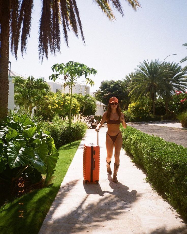 a woman walking down a sidewalk with a piece of luggage in her hand and palm trees behind her