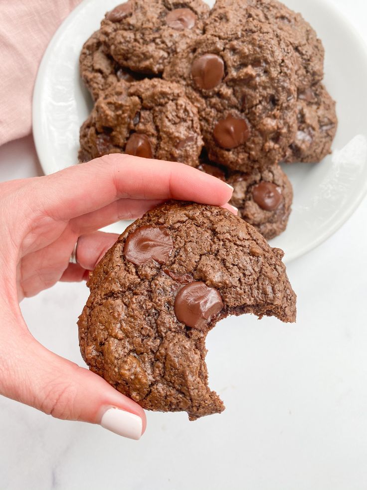 a hand holding a chocolate cookie in front of two other cookies on a white plate