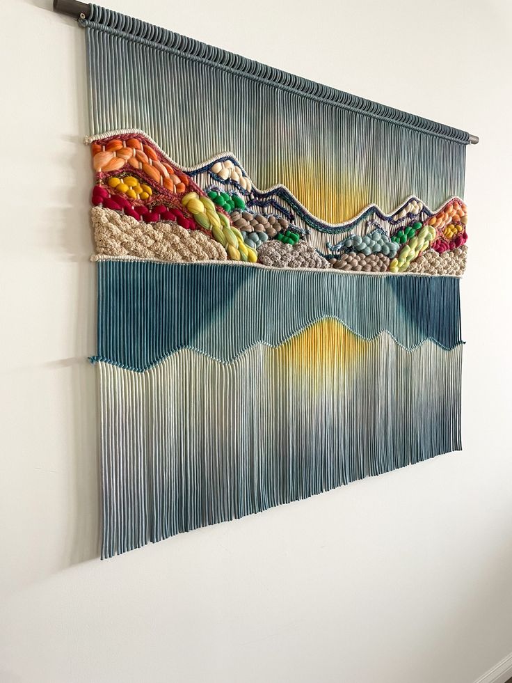 the wall hanging is decorated with multicolored yarns and fringes, including mountains