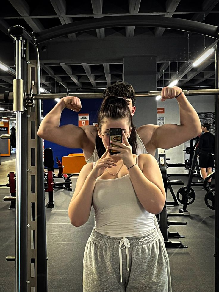 a woman taking a selfie with her cell phone in a gym area while standing next to a bar