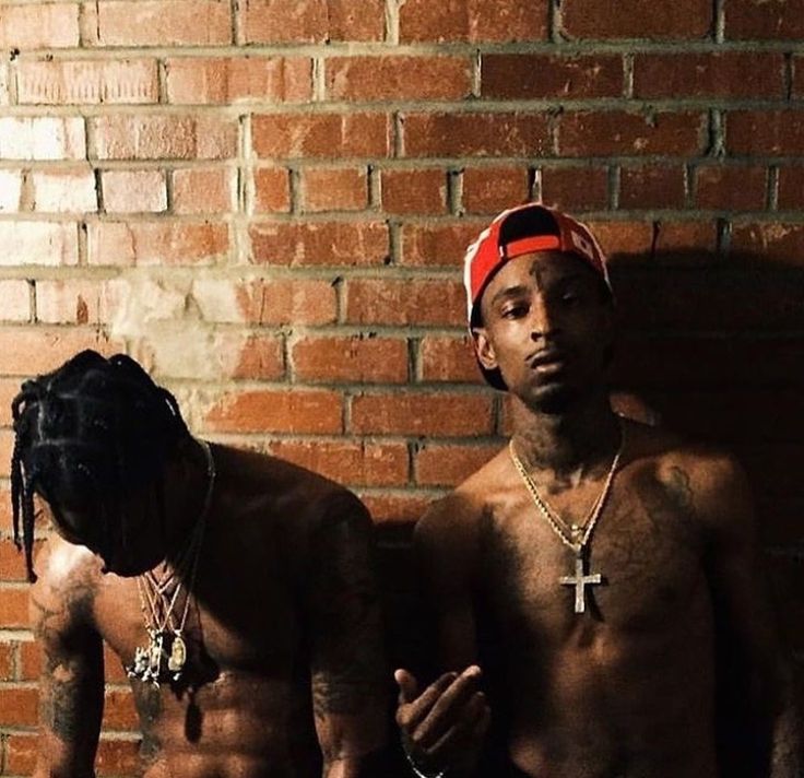 two men standing next to each other in front of a brick wall with one wearing a red bandana